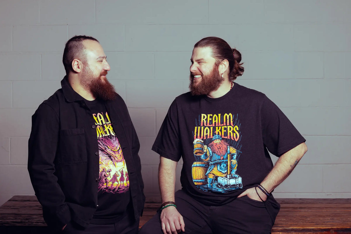 Two men wearing black t-shirts with colorful graphics and text, sitting on a wooden bench against a gray wall.