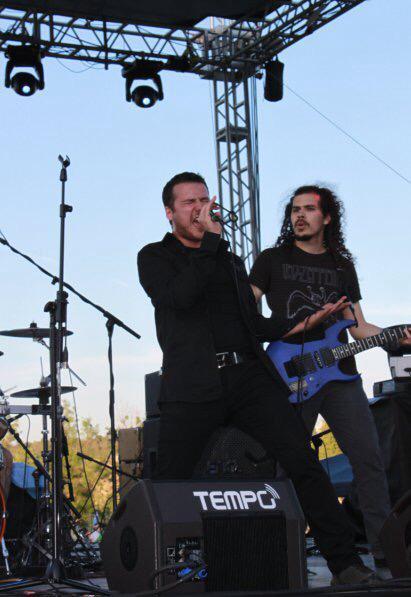 Two musicians performing on stage with musical equipment and a clear sky.